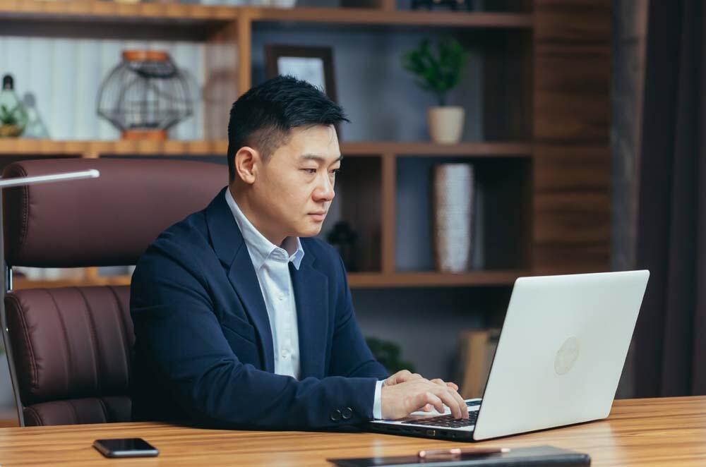 Asian man focused on his laptop while working in a modern office environment.